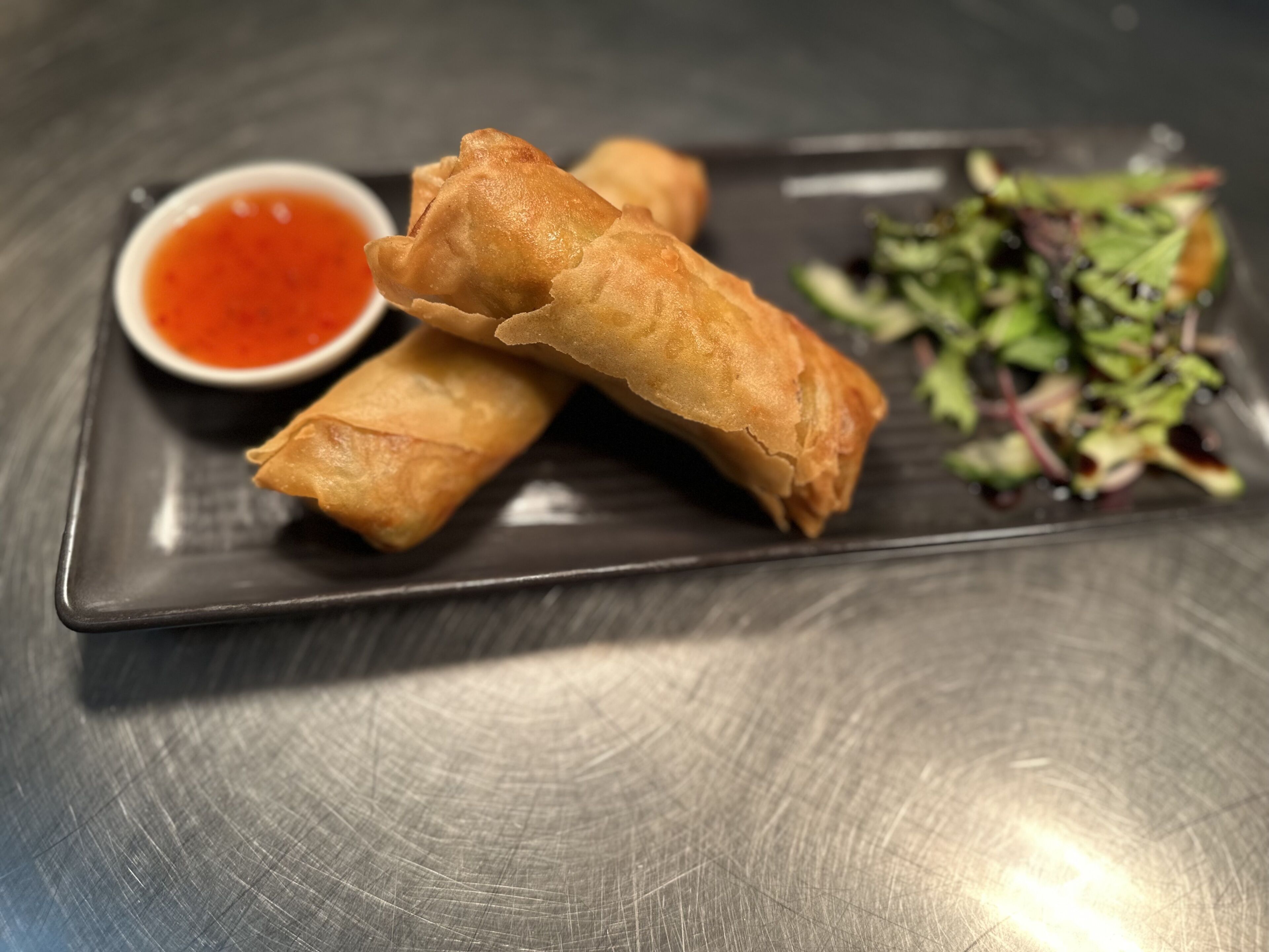 A plate with three spring rolls, a small bowl of red sauce, and a side salad.