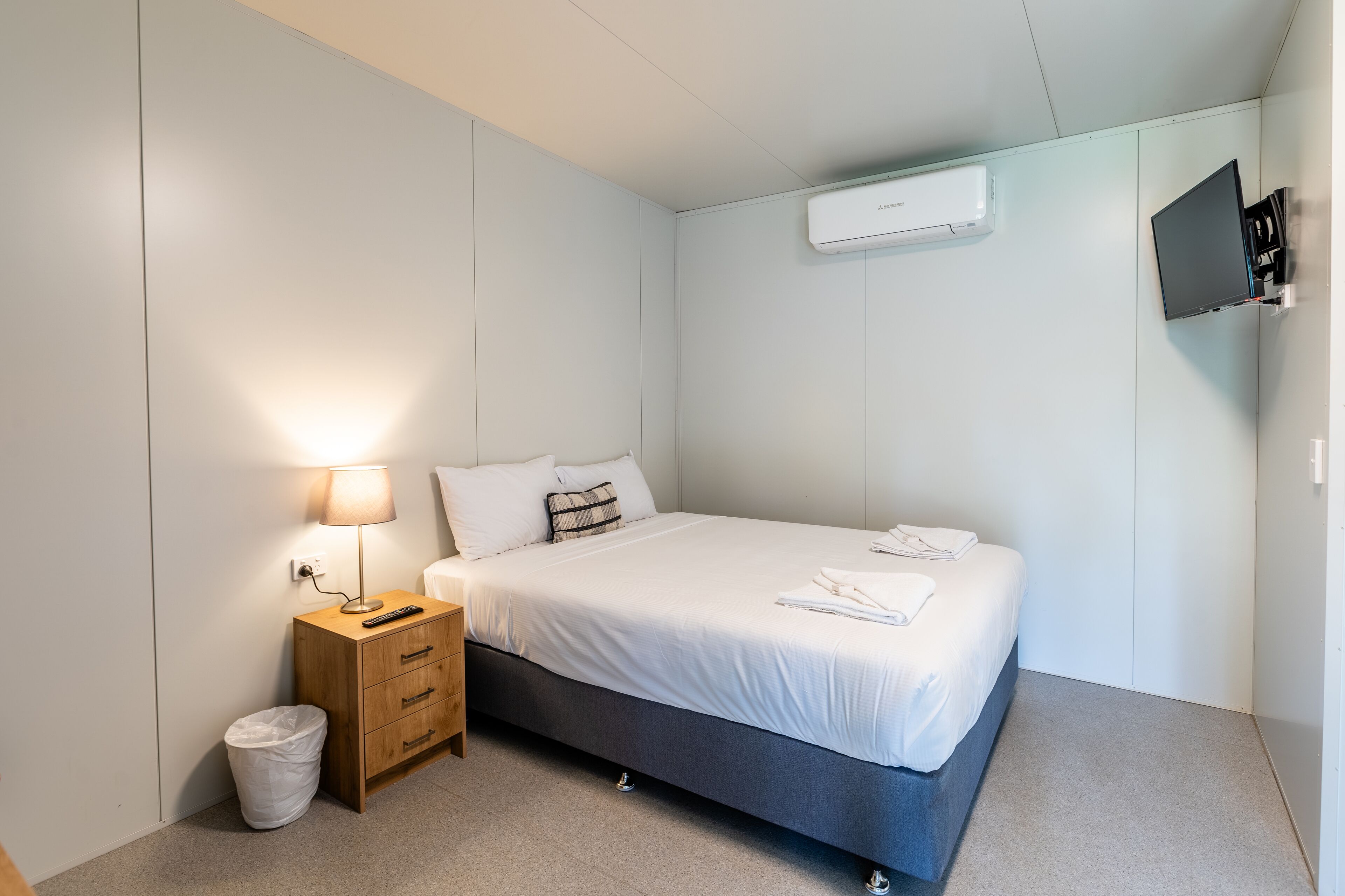 A modern bedroom with a bed, bedside lamp, wooden nightstand, and a TV mounted on the wall.