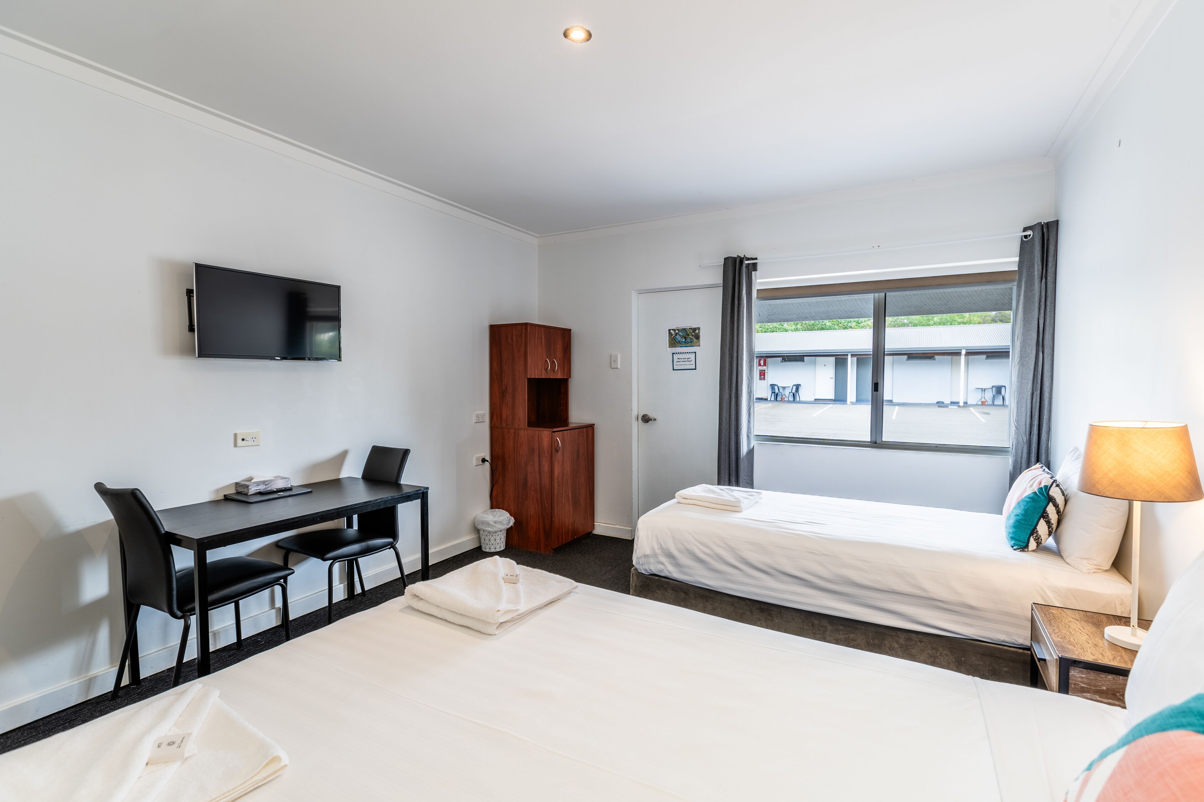 Une chambre d’hôtel comprenant un lit, un bureau avec des chaises, un téléviseur fixé au mur, une armoire et une fenêtre avec des rideaux.