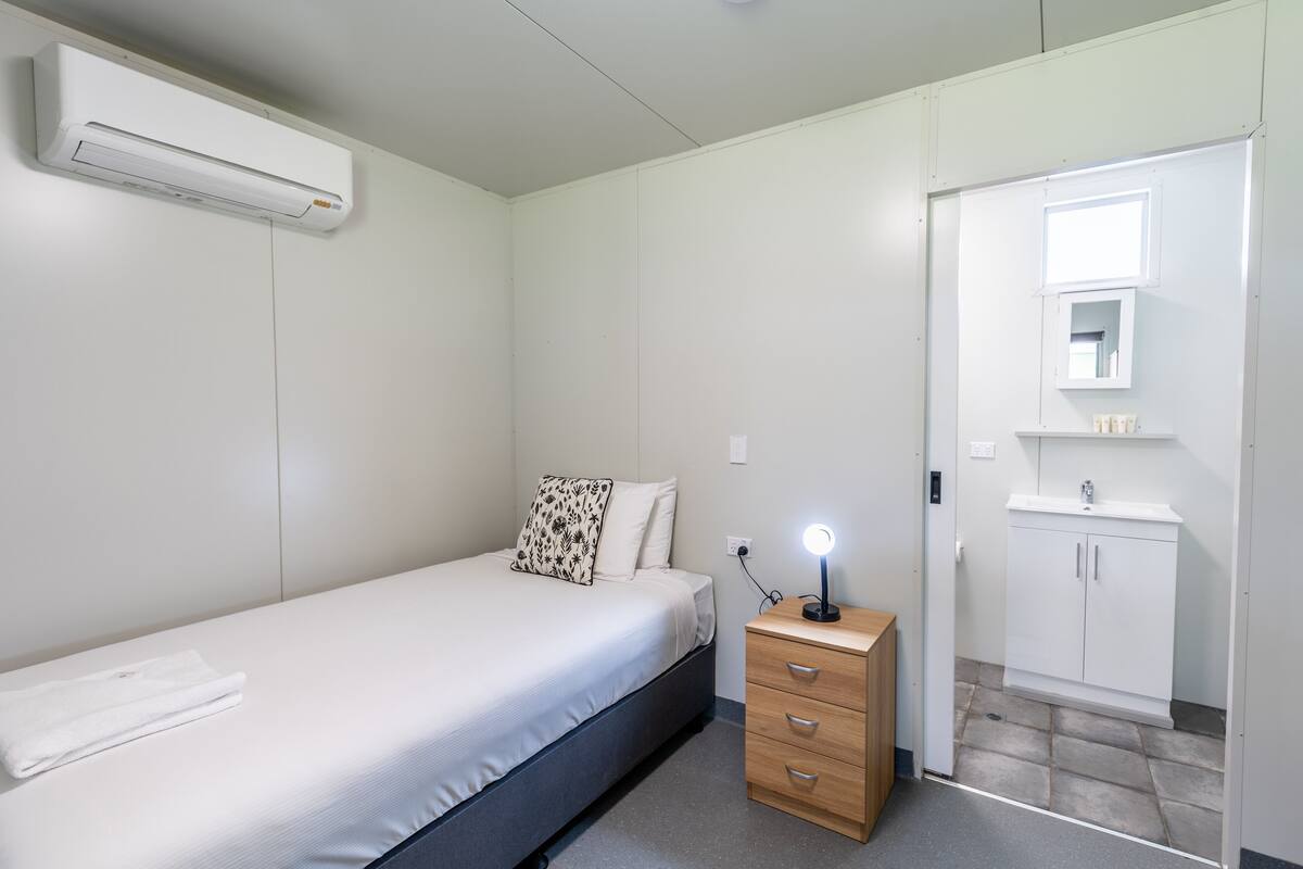 Une chambre à coucher comprenant un lit, une table de chevet avec une lampe, une armoire et une salle de bain avec un lavabo et un miroir.