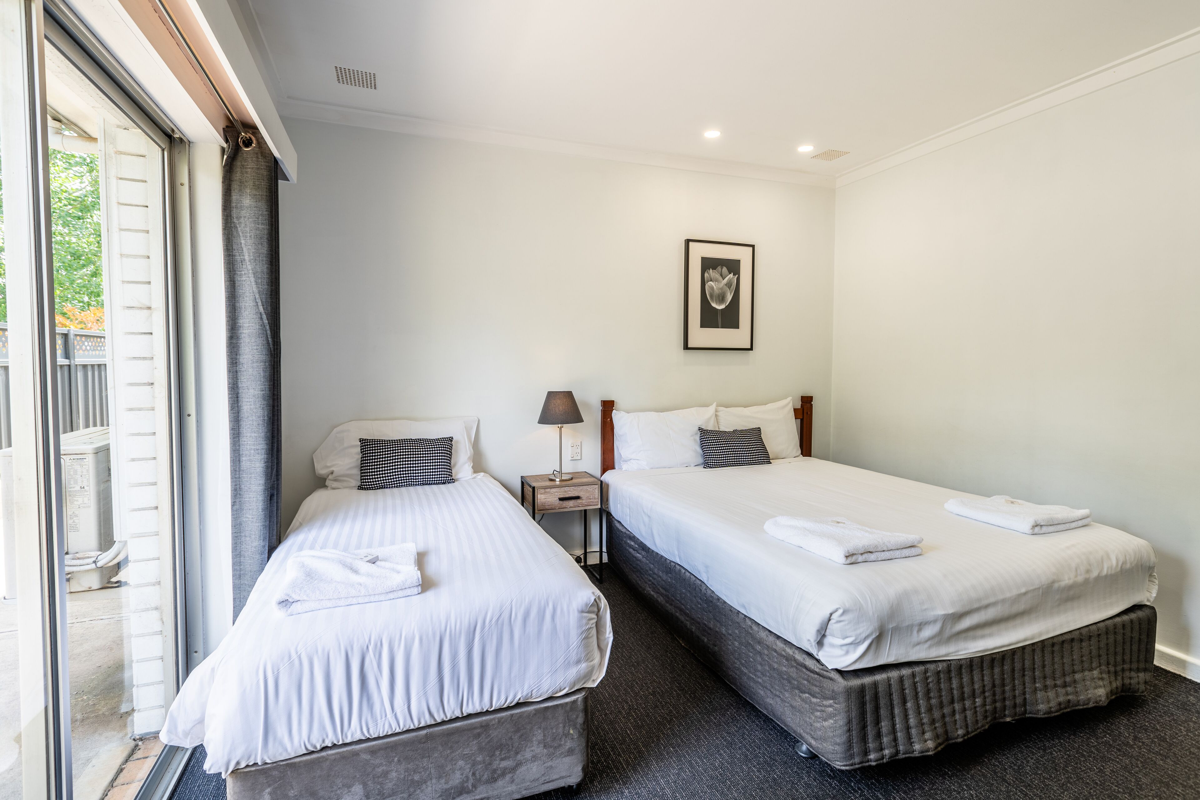 Una camera d'albergo con due letti, un comodino con una lampada, un quadro appeso alla parete e una porta vetrata scorrevole che dà su uno spazio all'aperto.
