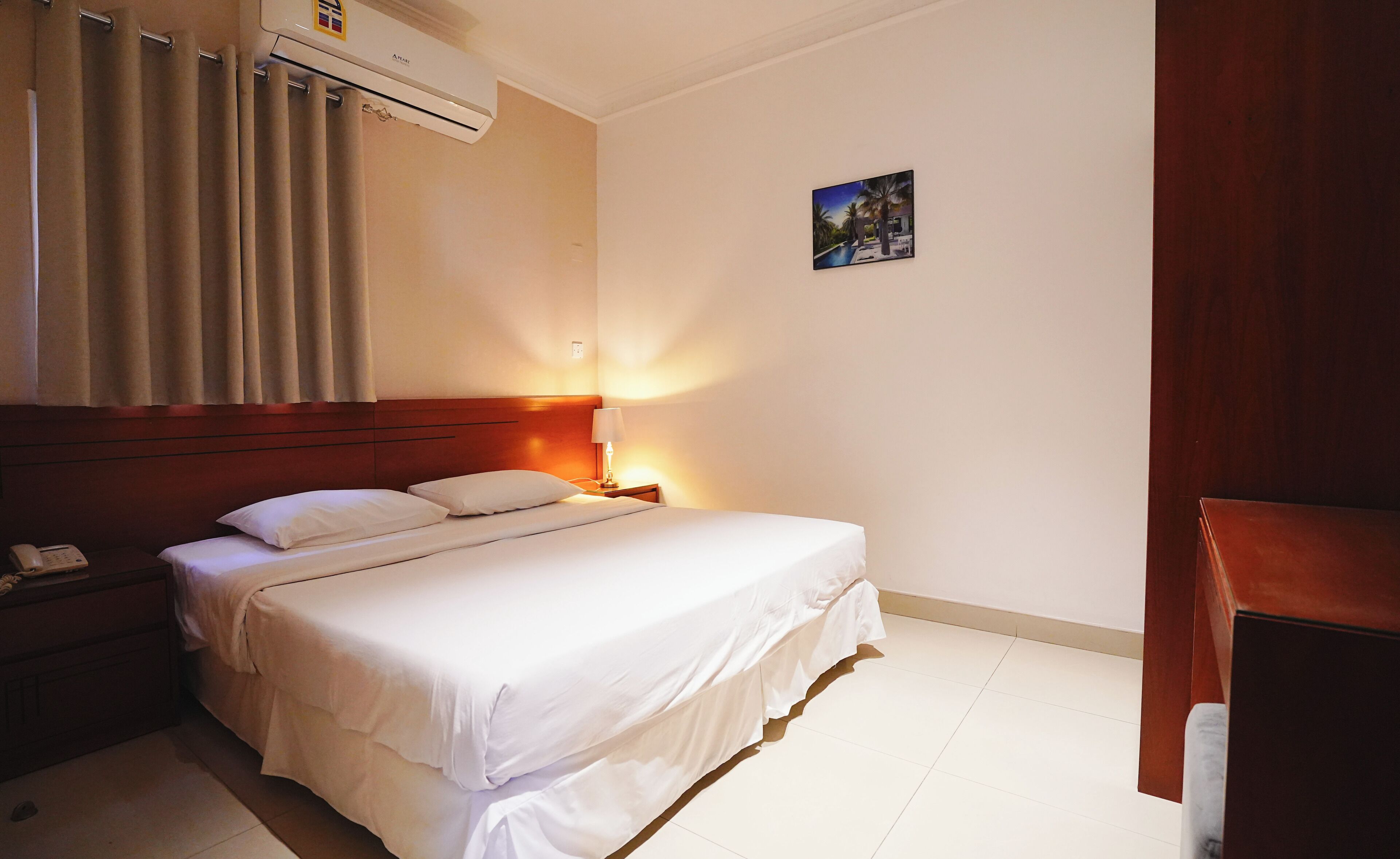 A hotel room with a bed, a bedside table with a lamp, a telephone, and an air conditioning unit.