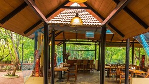 An outdoor covered dining area with stone flooring, wooden tables and chairs, and a central hanging light fixture. - The Windflower Jungle Resorts and Spa (Gundlupet)
