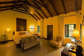 Quarto de hotel com cama, sofá, cadeira, mesa com luminária e janela com cortinas.