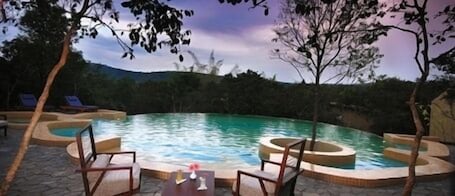 A circular pool with two lounge chairs and a small table, surrounded by trees and a mountainous backdrop.