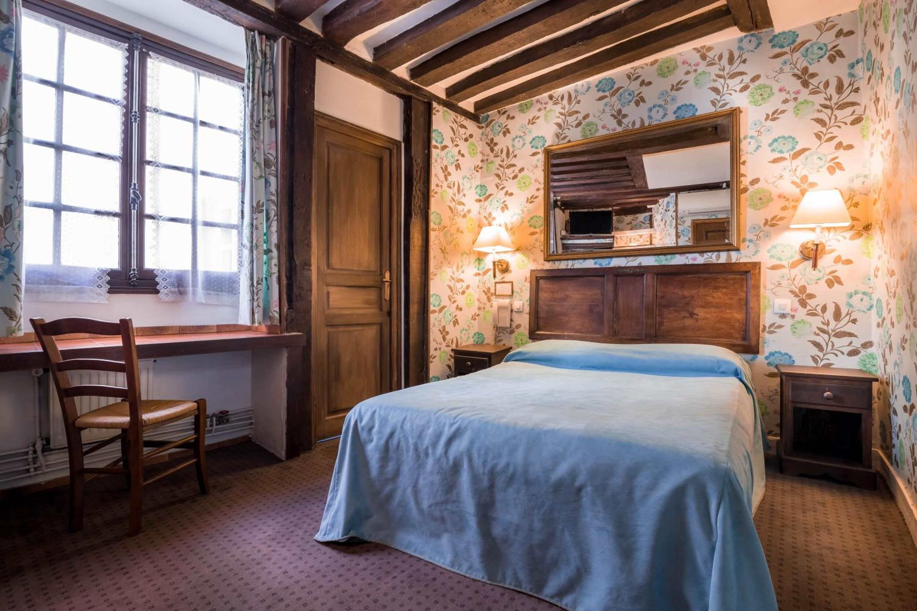 Een slaapkamer met een bed, een houten bureau, een stoel, een raam en een spiegel.