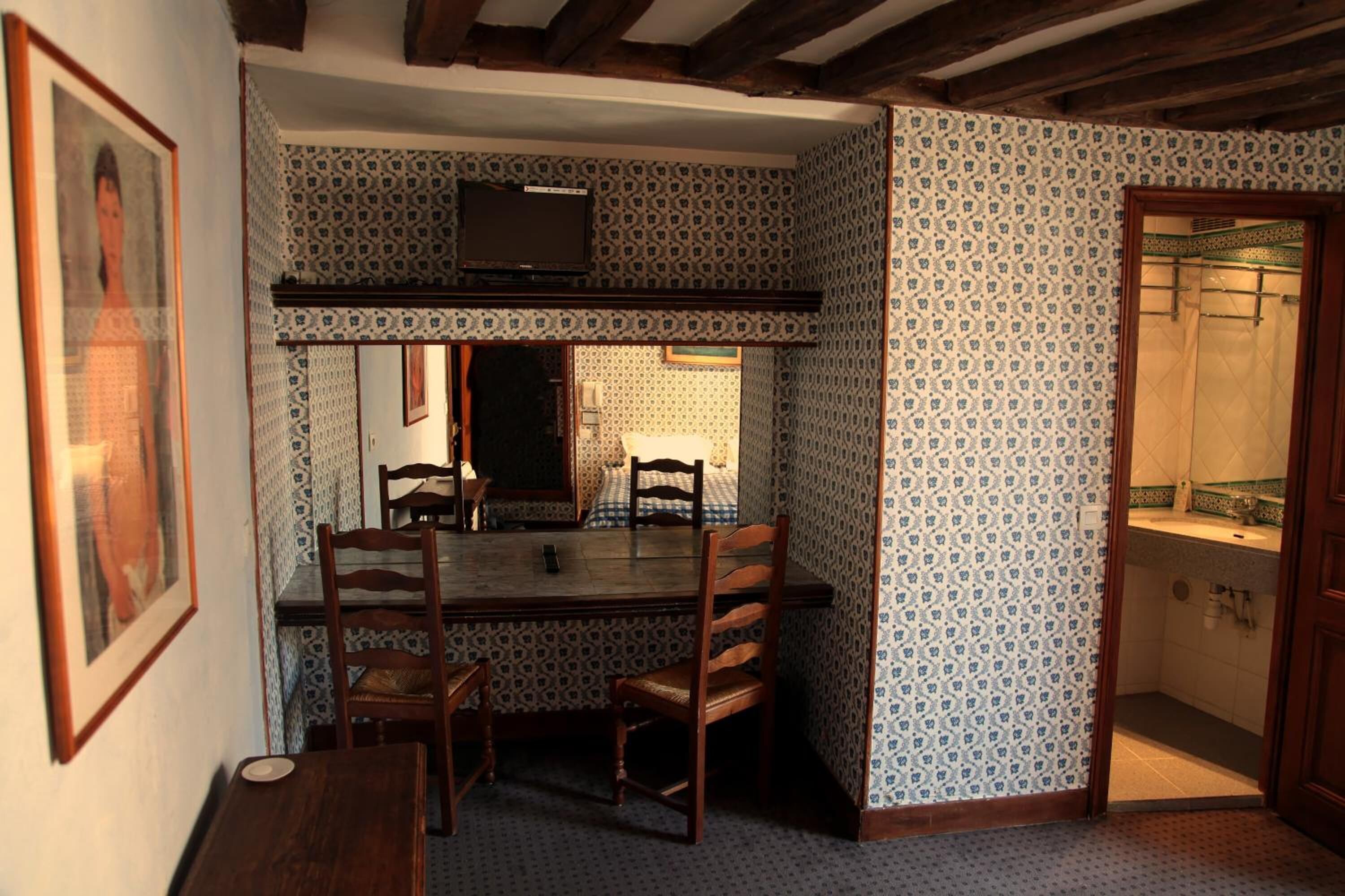 Un coin repas avec une table et des chaises en bois, un téléviseur placé au-dessus de la table, un papier peint à motifs et une petite salle de bain visible à travers une porte ouverte.