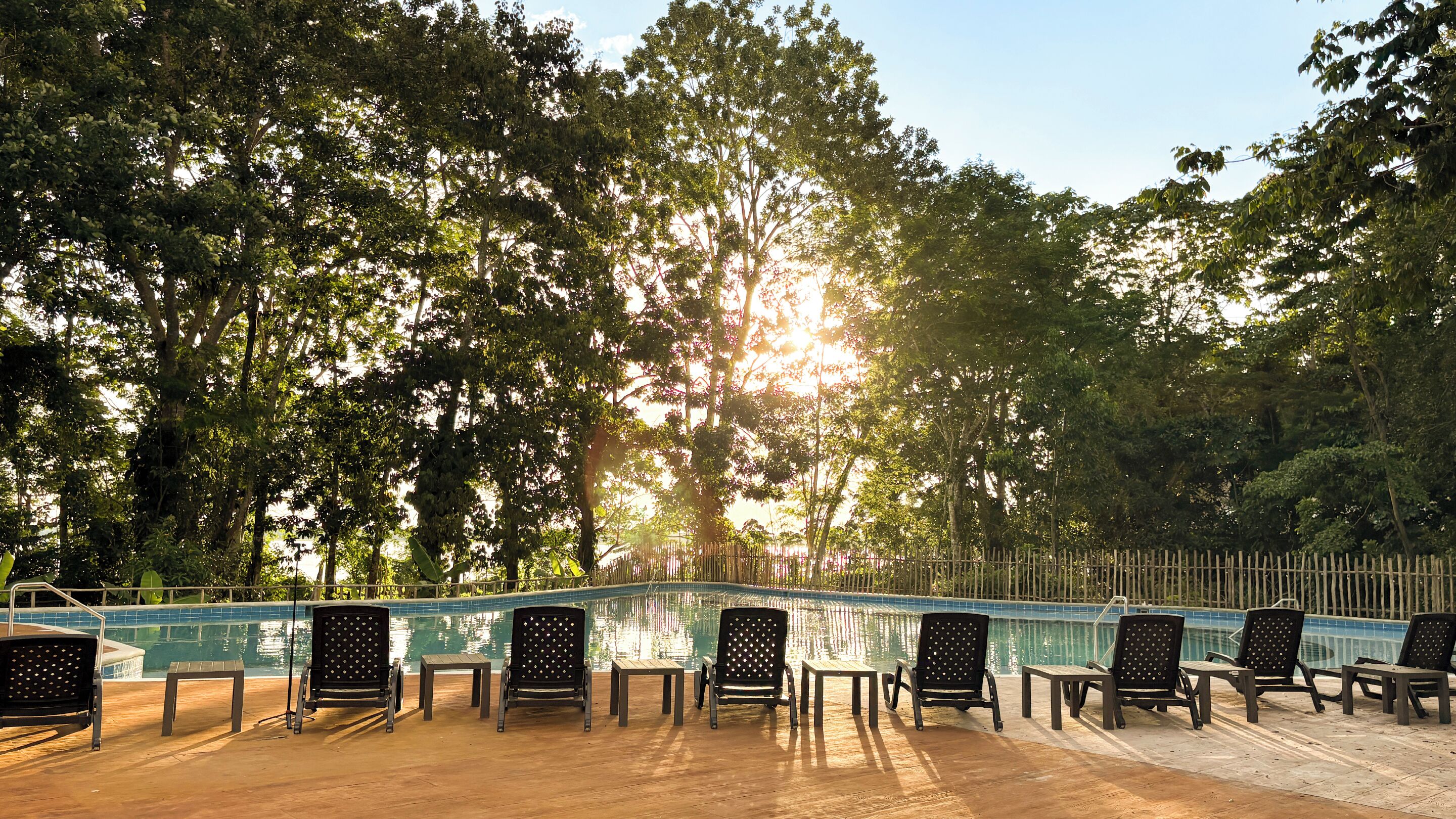 A poolside area with lounge chairs and tables, surrounded by trees and a wooden fence.