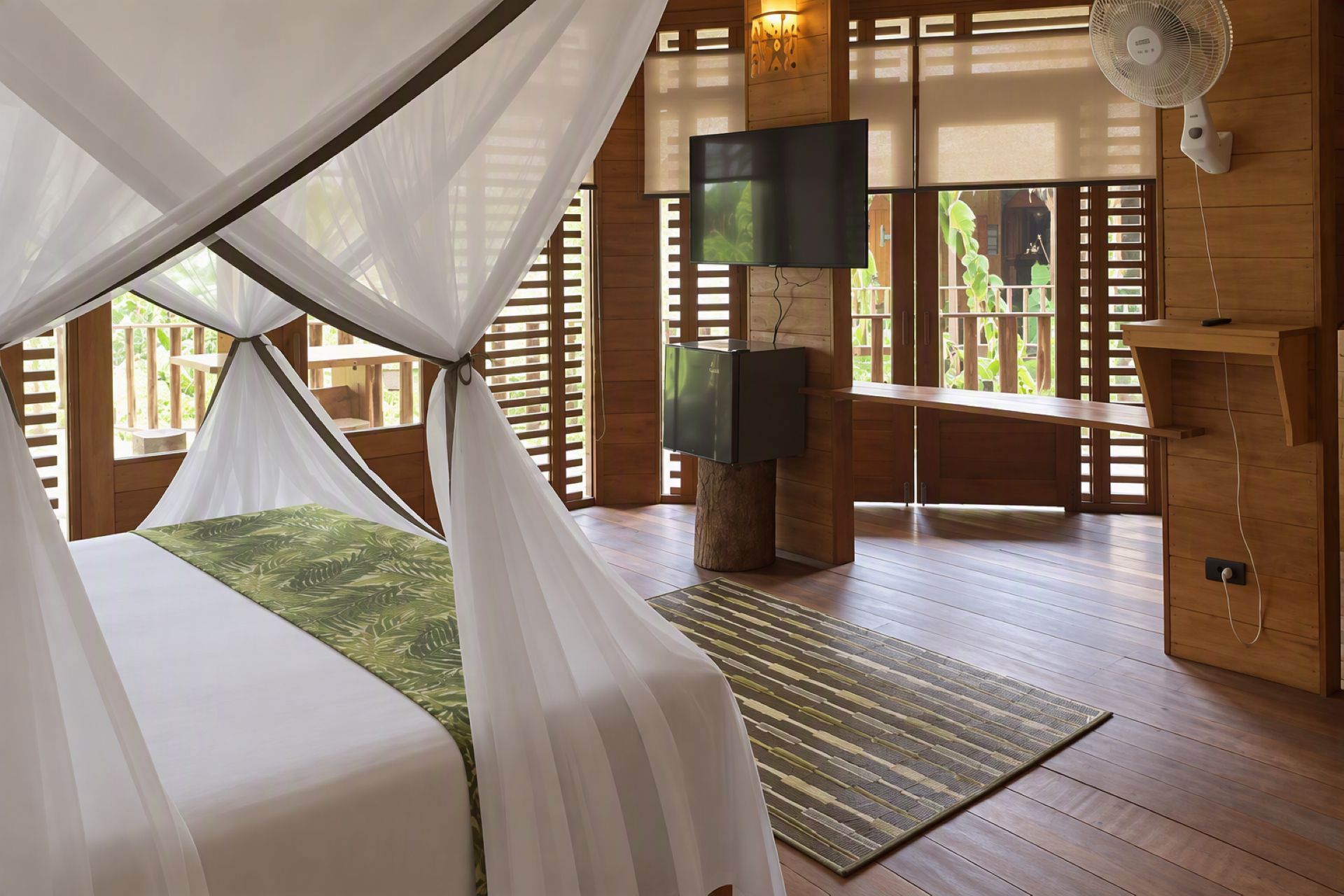 A tented room with a bed, a flat-screen TV, a fan, and a wooden desk.