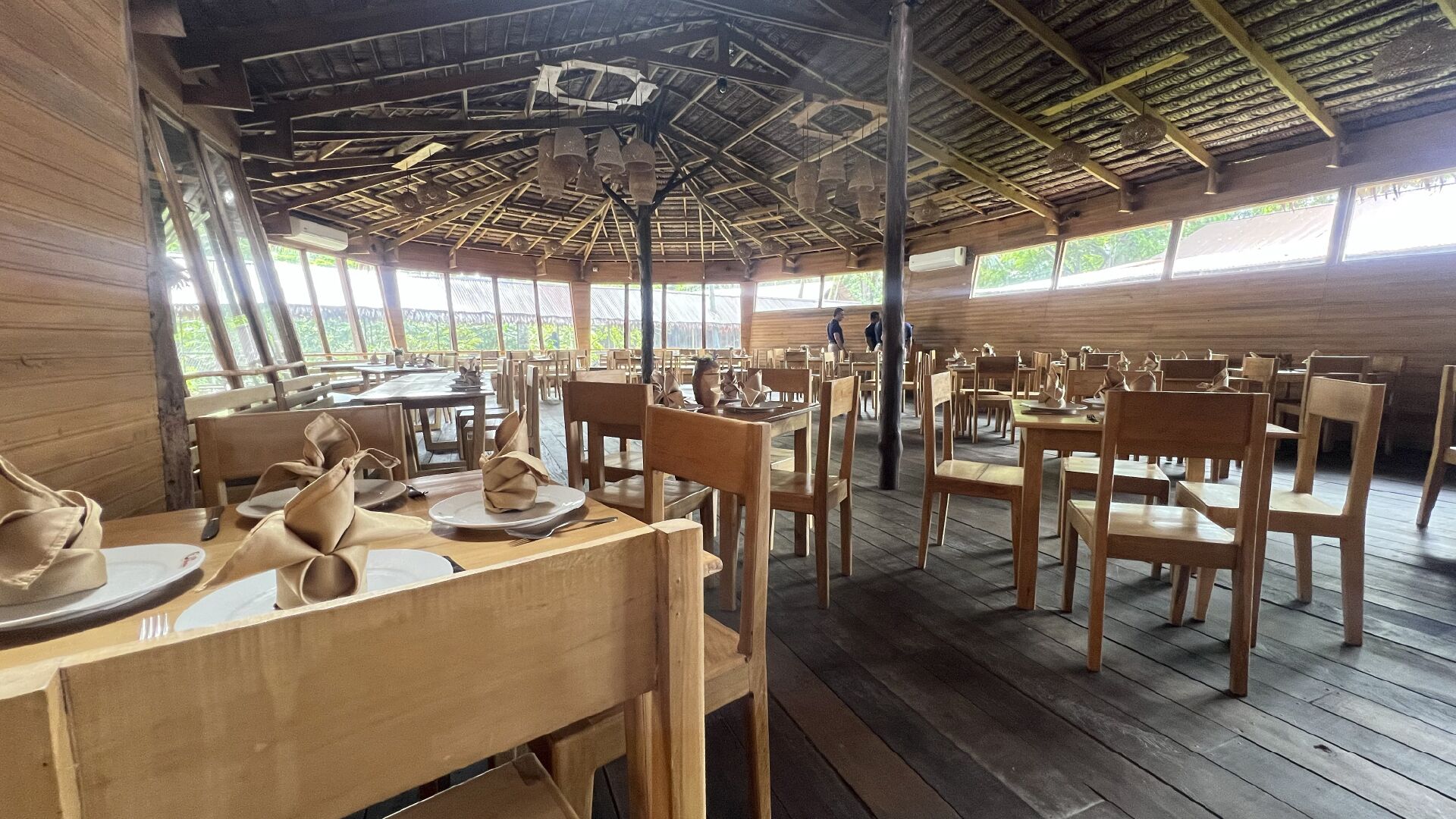 Un vaste espace dédié à la salle à manger, avec des tables et des chaises en bois, de grandes fenêtres et un plafond haut avec des poutres apparentes.