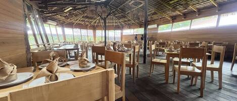 Un vaste espace dédié à la salle à manger, avec des tables et des chaises en bois, de grandes fenêtres et un plafond haut avec des poutres apparentes.