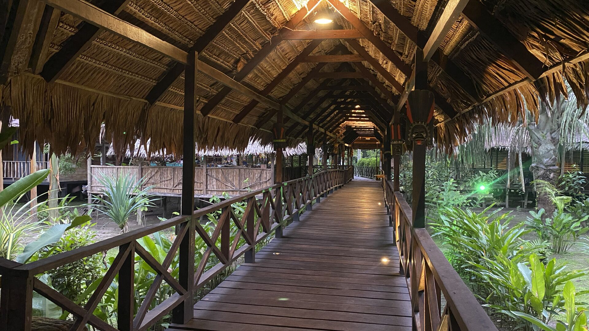 A wooden walkway with a thatched roof, surrounded by lush greenery.