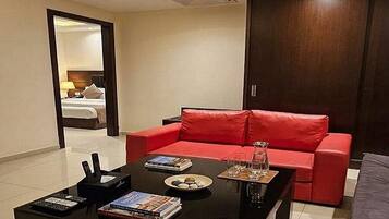 Una sala moderna con un sofá rojo, una mesa de centro negra con libros y controles remotos, y vista a un dormitorio por una puerta abierta.