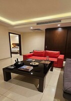 Una sala moderna con un sofá rojo, una mesa de centro negra con libros y controles remotos, y vista a un dormitorio por una puerta abierta.