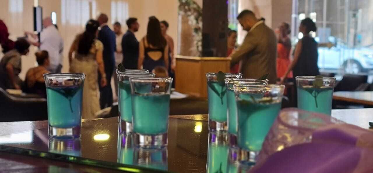 Zona de bar con varios vasos de una bebida azul, una barra y gente al fondo.