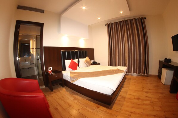 A hotel room with a wooden floor, a bed with white linens, a red chair, a nightstand, a desk, and a television on the wall.