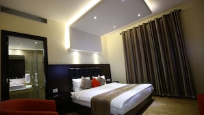 Una habitación de hotel moderna con una cama grande, mesitas de noche, un espejo, una silla roja y un baño con lavabo y ducha.