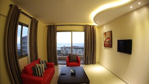 Una sala moderna con un sofá rojo, una mesa de centro negra y un sillón rojo. Se ve un ventanal con cortinas, un televisor de pantalla plana montado en la pared y un cuadro sobre el televisor.
