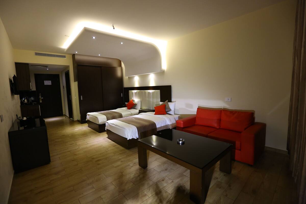 Une chambre d’hôtel moderne avec un plancher en bois, deux lits simples, un canapé rouge, une table basse noire et un grand miroir au-dessus du canapé.