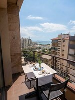 Un balcón con una mesa lista para dos personas, con vista a un paisaje urbano que incluye edificios y una masa de agua.