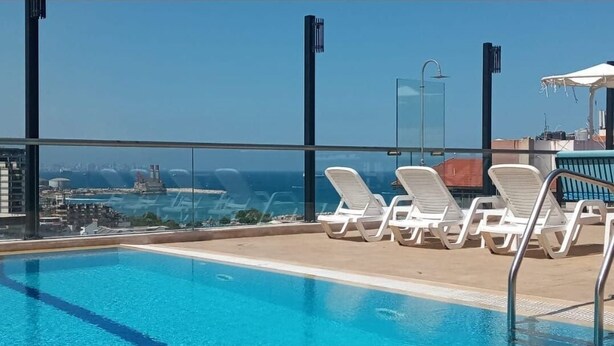 Una piscina en la azotea con agua azul cristalina, sillas de descanso blancas y vistas a la ciudad y al océano.