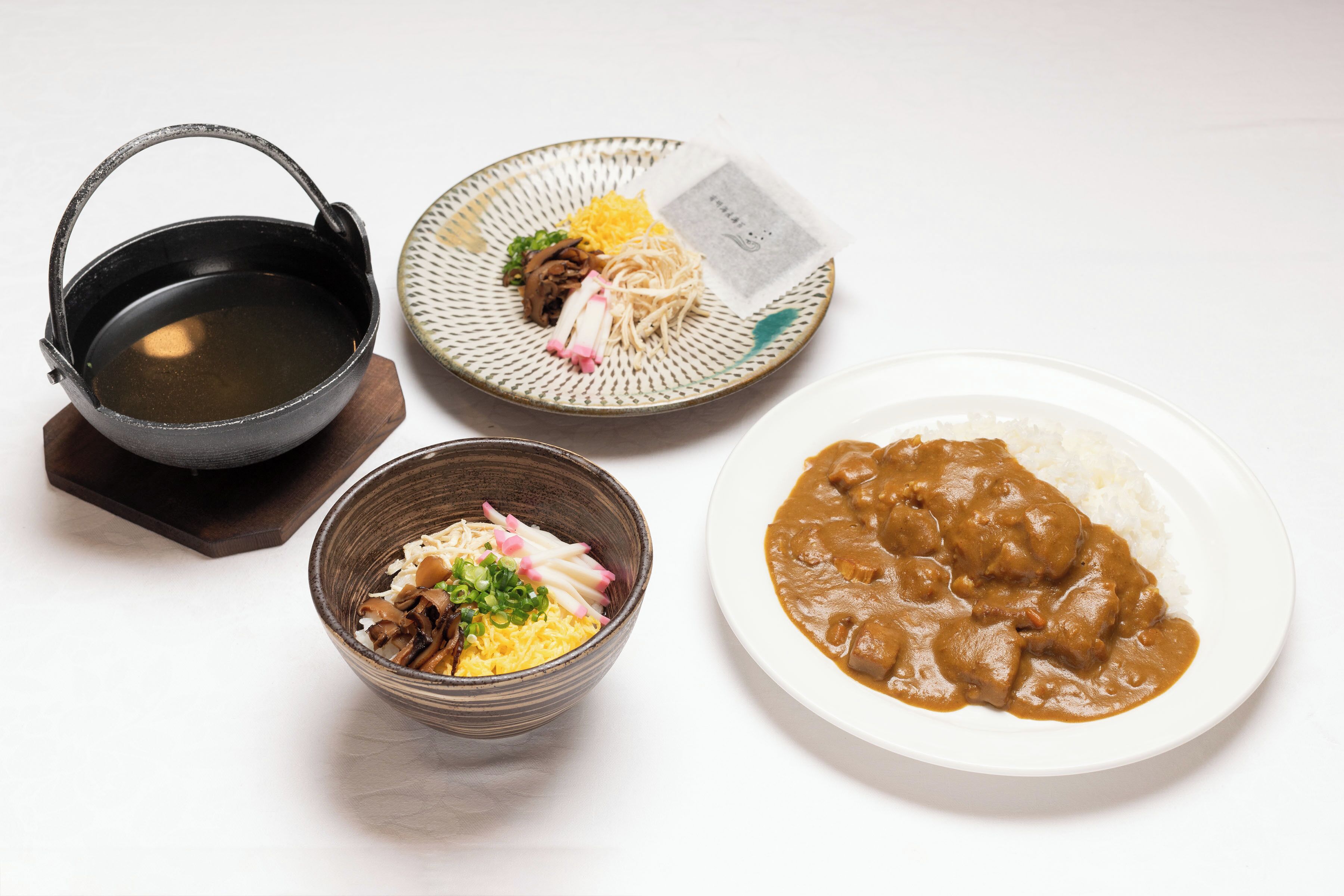 Una mesa con un tazón de sopa, un plato de arroz con curry, un bol de fideos con verduras y una pequeña olla.