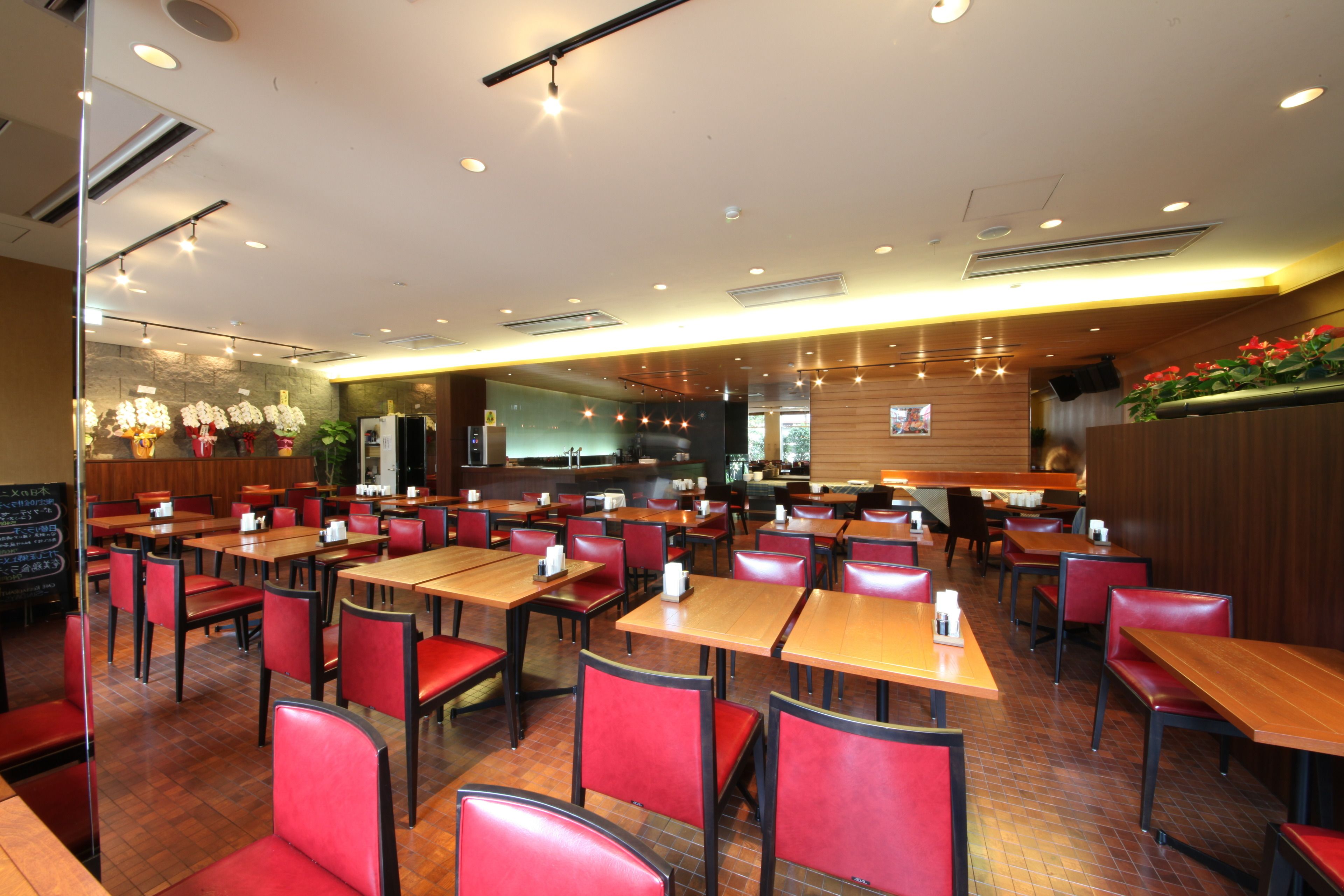Un restaurant avec plusieurs tables et des chaises rouges, un espace cuisine à l’arrière-plan et un comptoir en bois décoré de fleurs rouges.