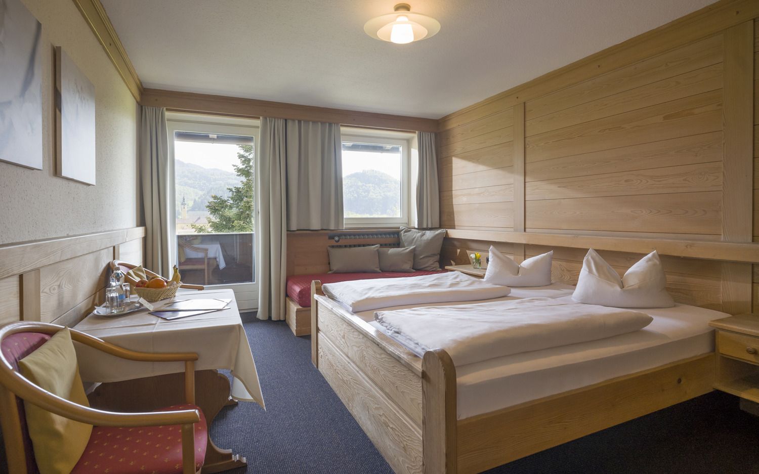 A hotel room with two beds, a desk with a chair, a small table with a basket of fruit, and a view of mountains through the window.