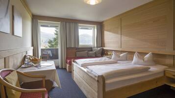 A hotel room with two beds, a desk with a chair, a small table with a basket of fruit, and a view of mountains through the window.