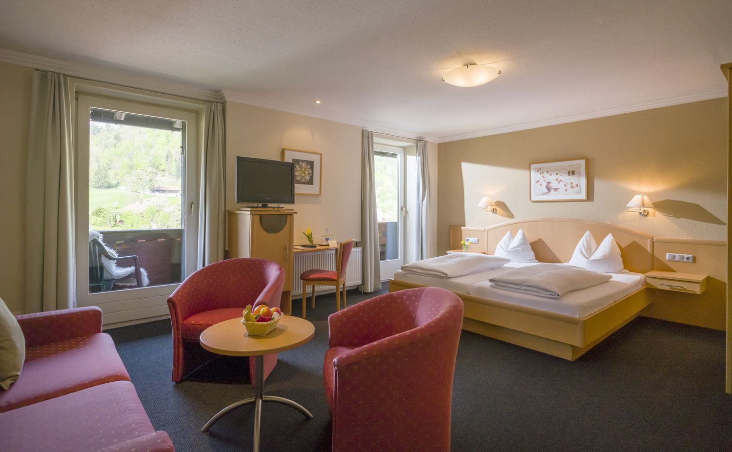 A hotel room with a bed, two red armchairs, a small table with fruit, a TV, and a window with curtains.
