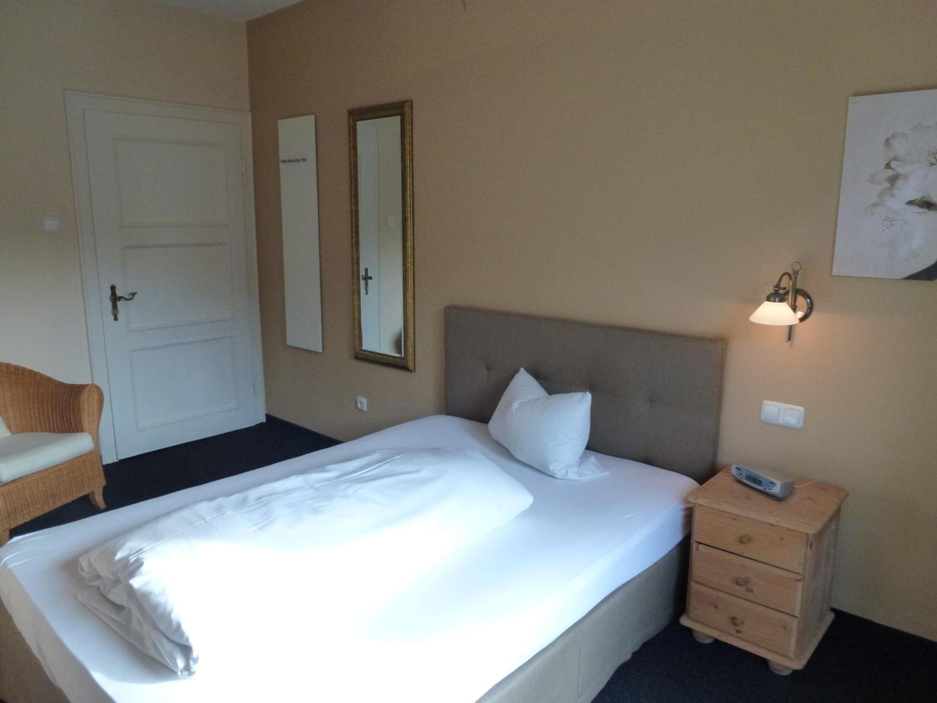 A hotel room with a bed, a bedside table with a lamp, a chair, and a door.