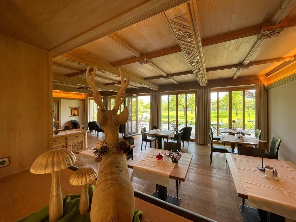 A dining area with tables and chairs, a large window with a view of greenery, and a ceiling with intricate woodwork.