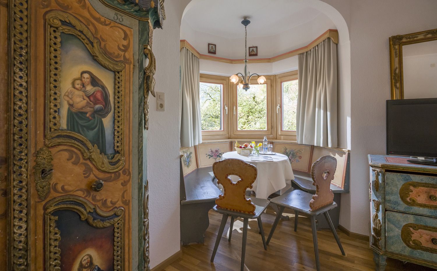 A dining area with a round table, chairs, and a chandelier. There is a window with curtains and a view of greenery outside.