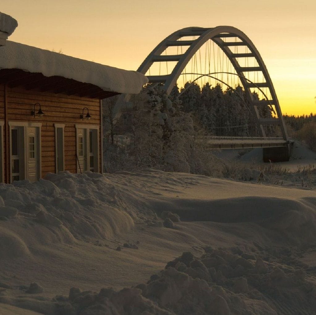 En stuga byggd av trä med ett snötäckt tak, en brokonstruktion av metall och en skog i bakgrunden.