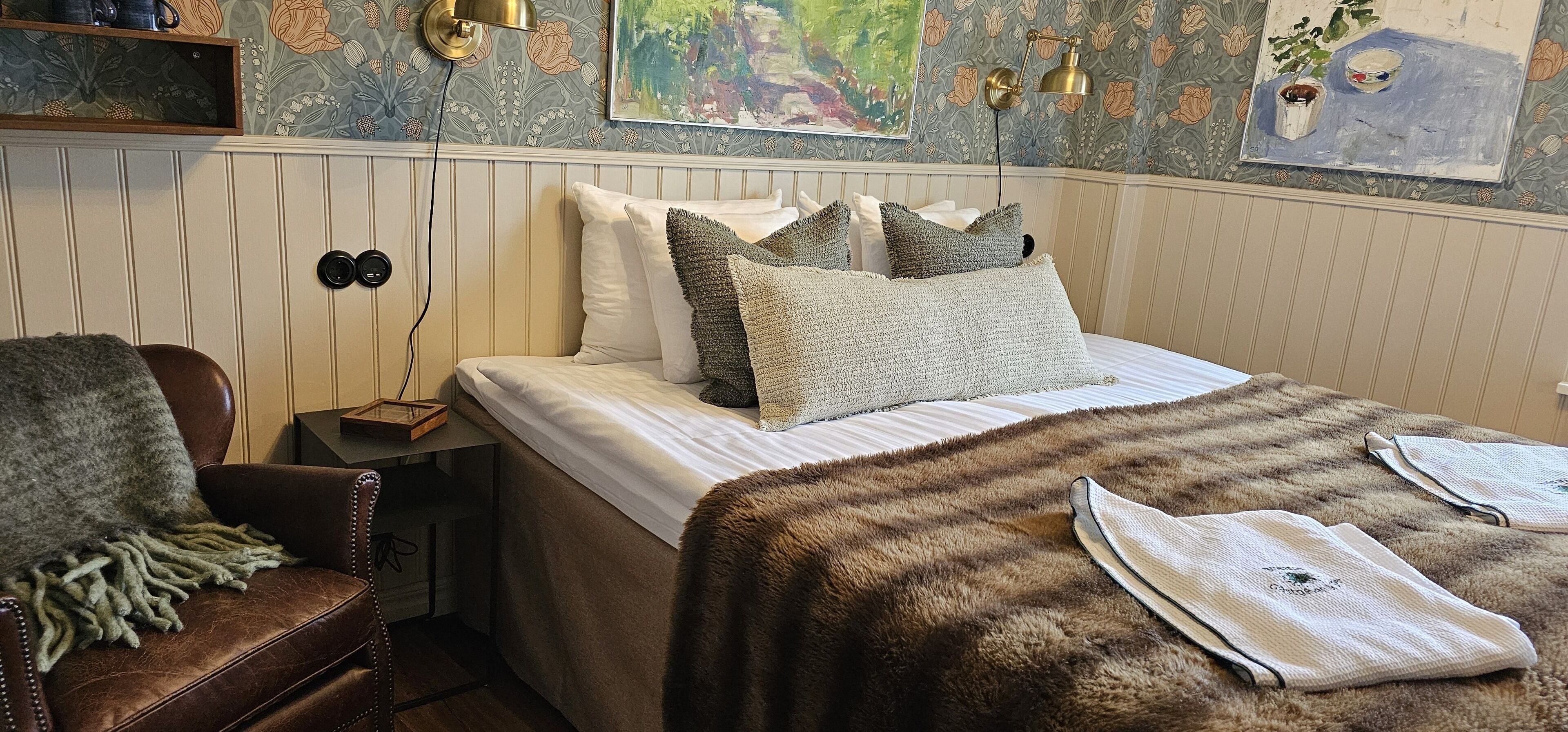 Un dormitorio con cama, una silla, una mesita de noche y un papel tapiz floral con obras de arte.