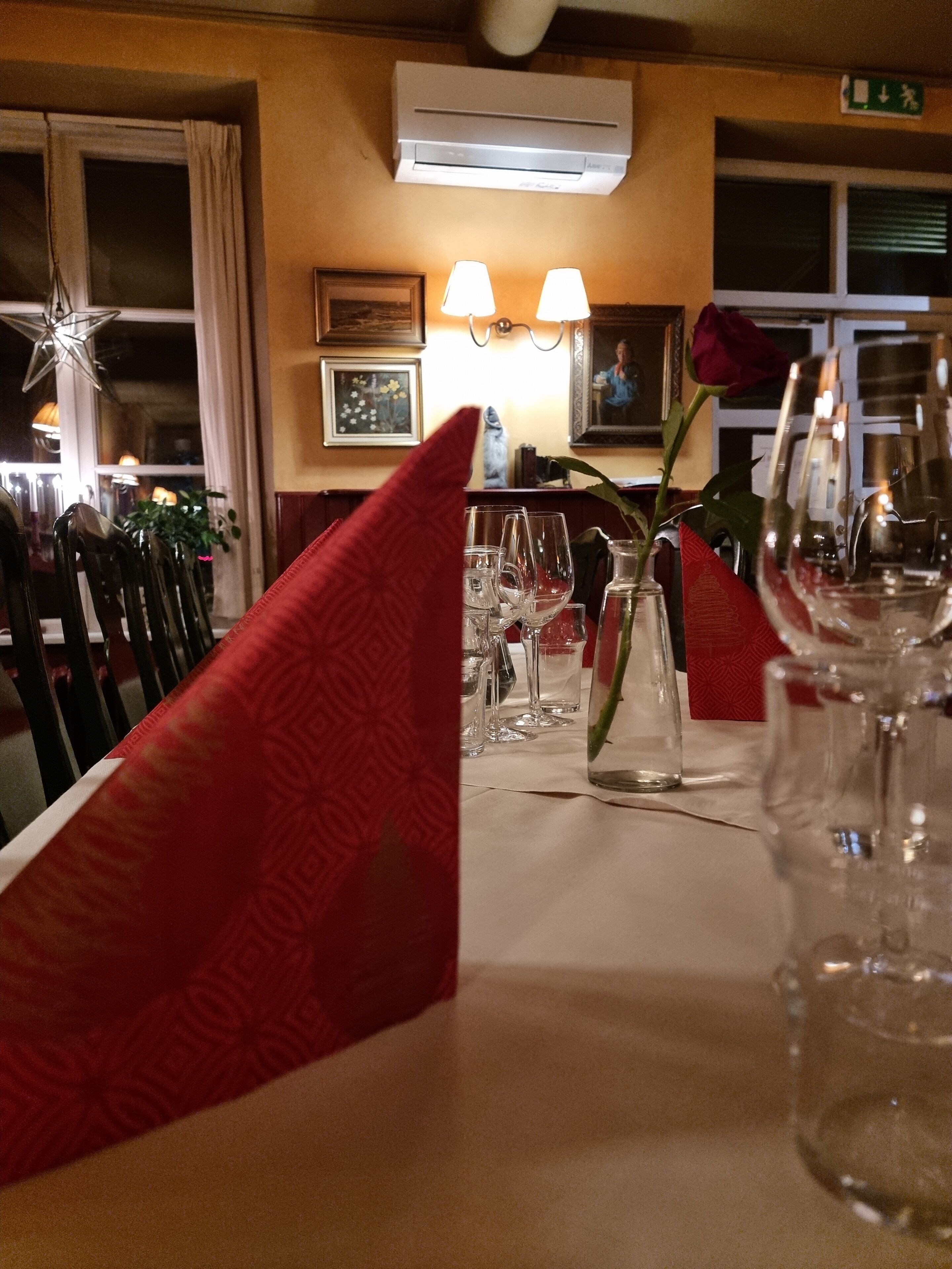 A restaurant interior with a table set for dining, chairs, a vase with a rose, and a wall-mounted air conditioner.