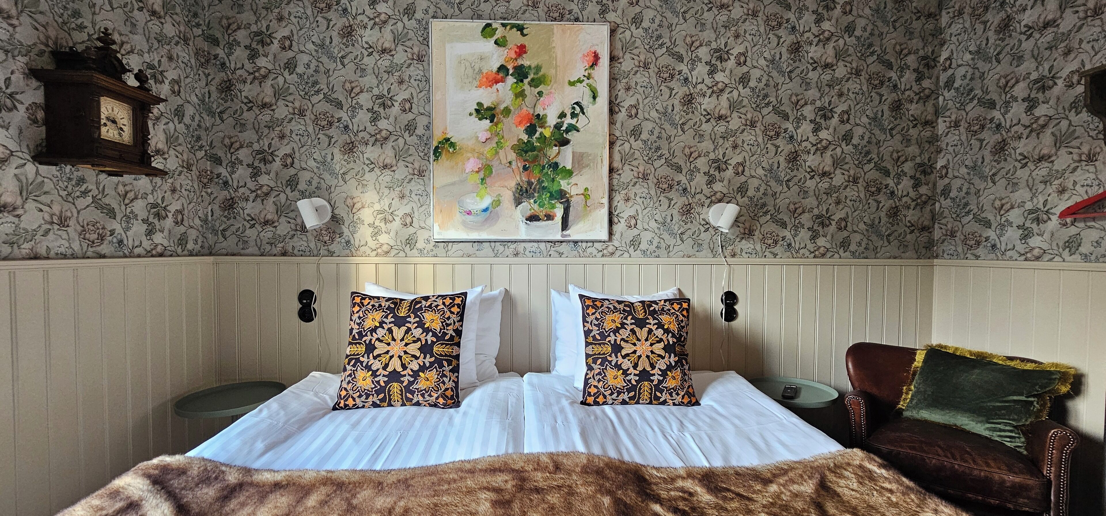 A bedroom with a bed, two decorative pillows, a painting of flowers, a clock, and a chair.
