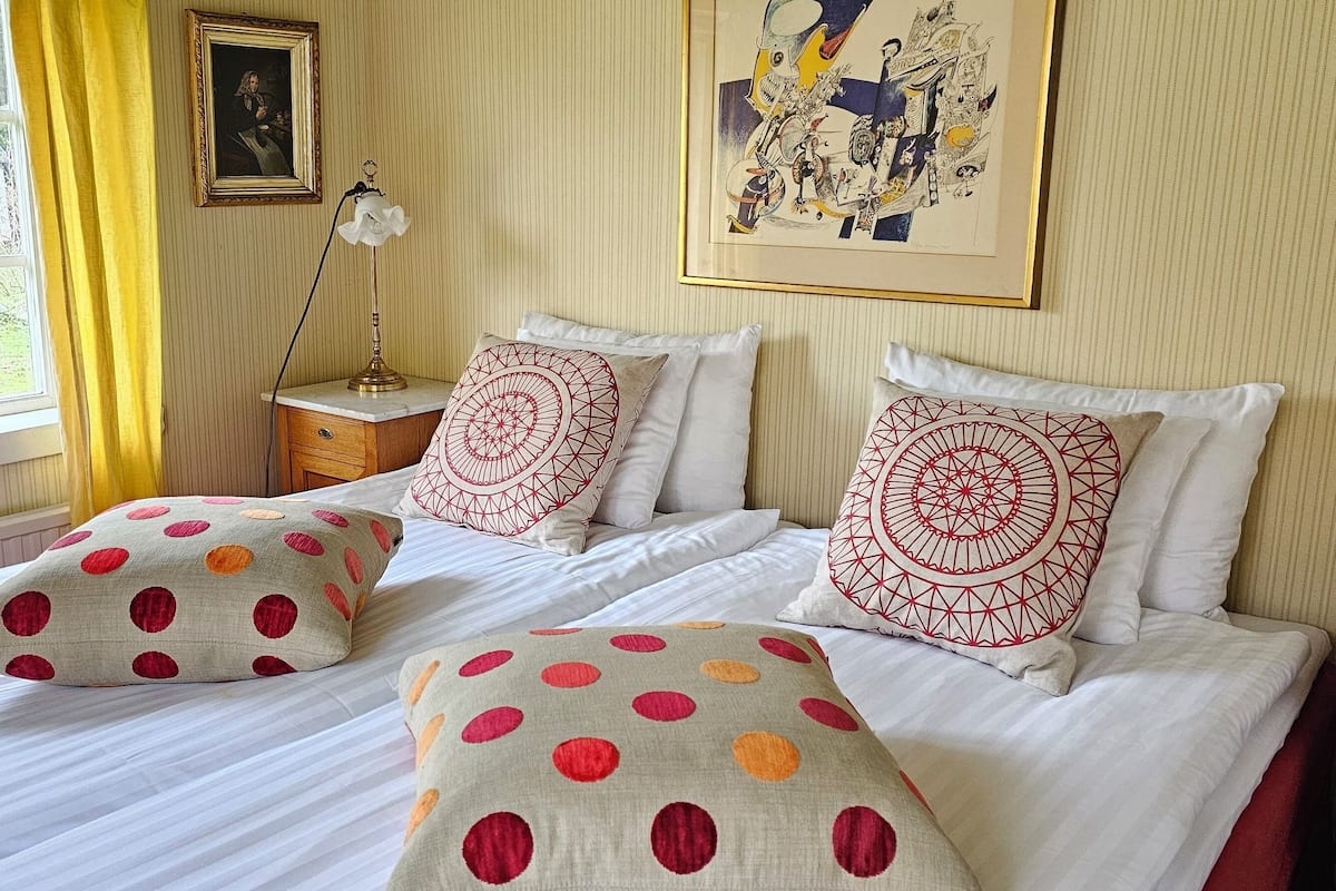 A bedroom with two beds, each with patterned pillows, a bedside table with a lamp, and a window with curtains.