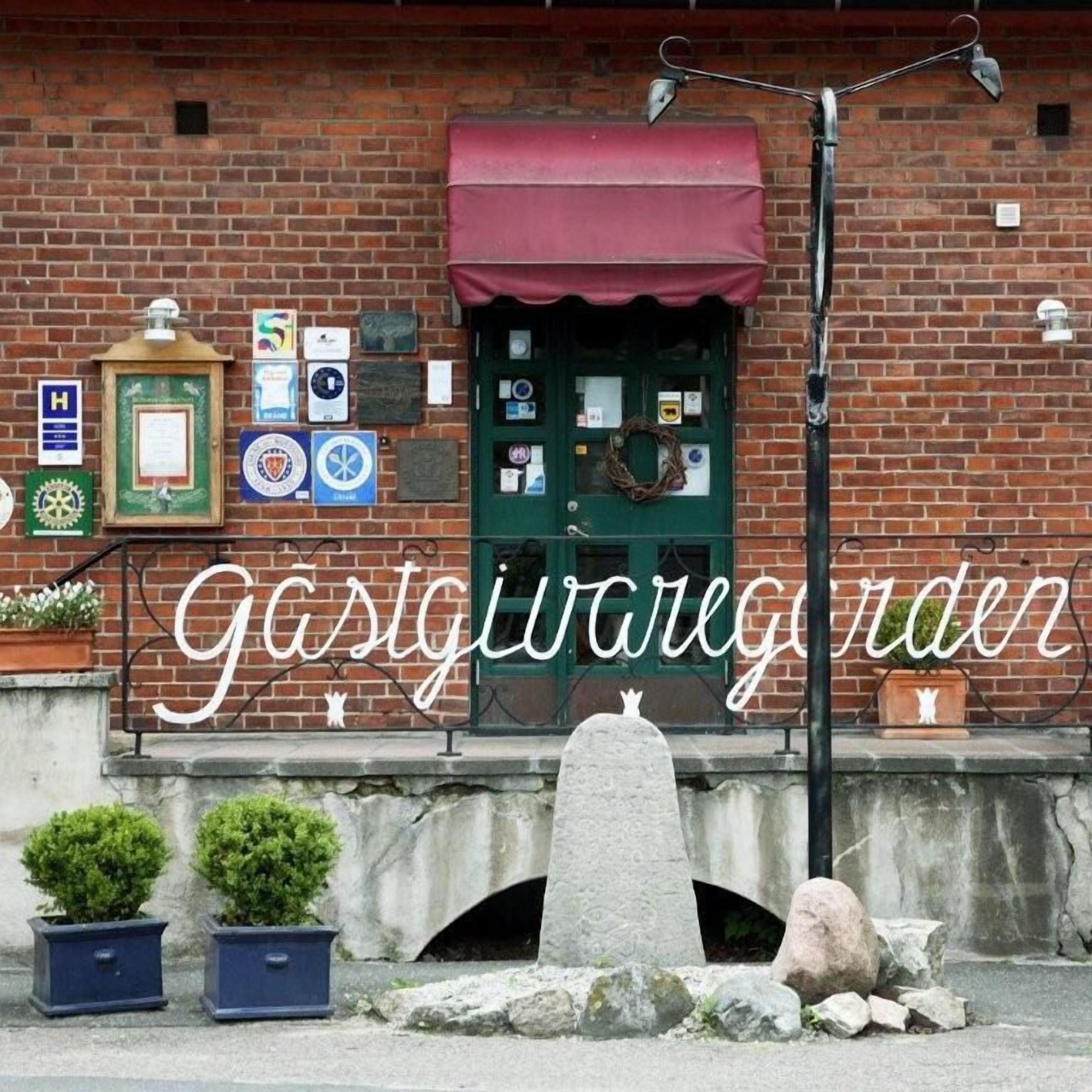 En byggnad av tegel med en grön dörr, ett rött markis och flera plaketter på väggen. Framför byggnaden står två krukväxter.