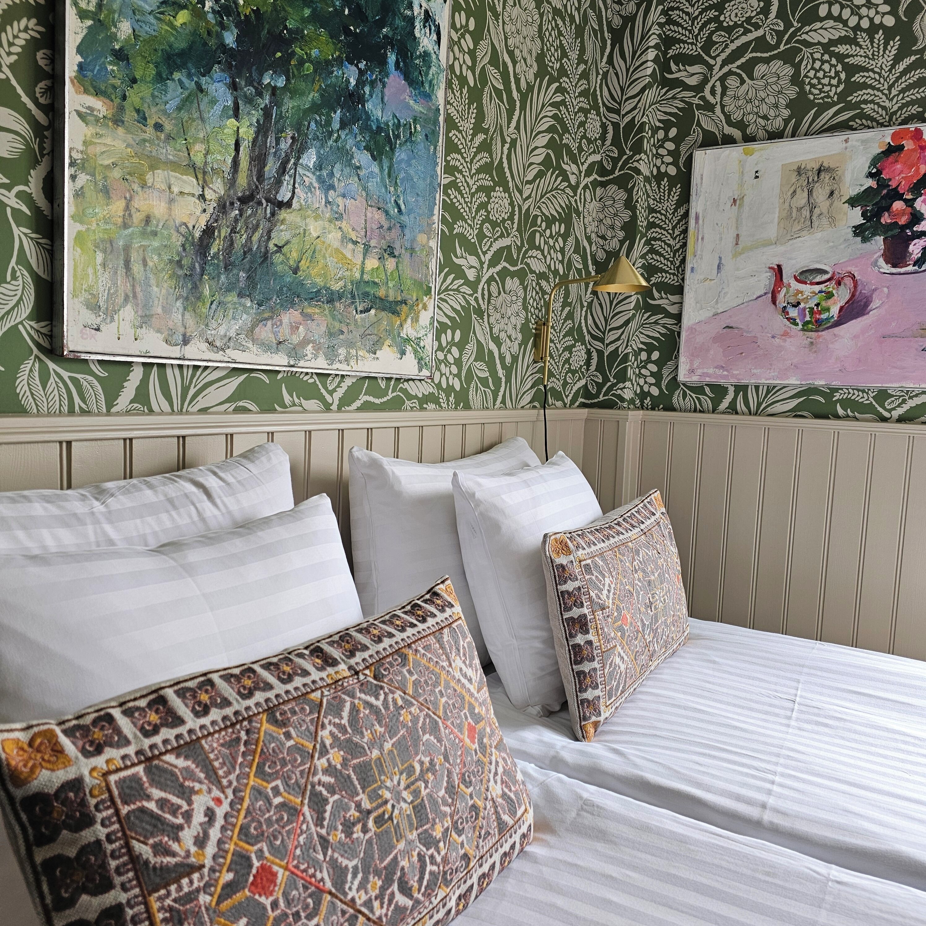 A neatly made bed with white linens and patterned pillows, a green floral wallpaper, and a painting of a forest scene.