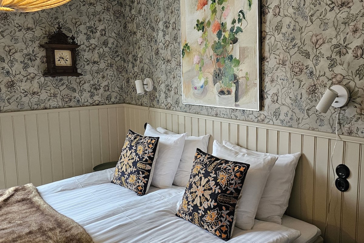 A neatly made bed with white linens and floral-patterned pillows, a wall clock, and a floral painting above the bed.
