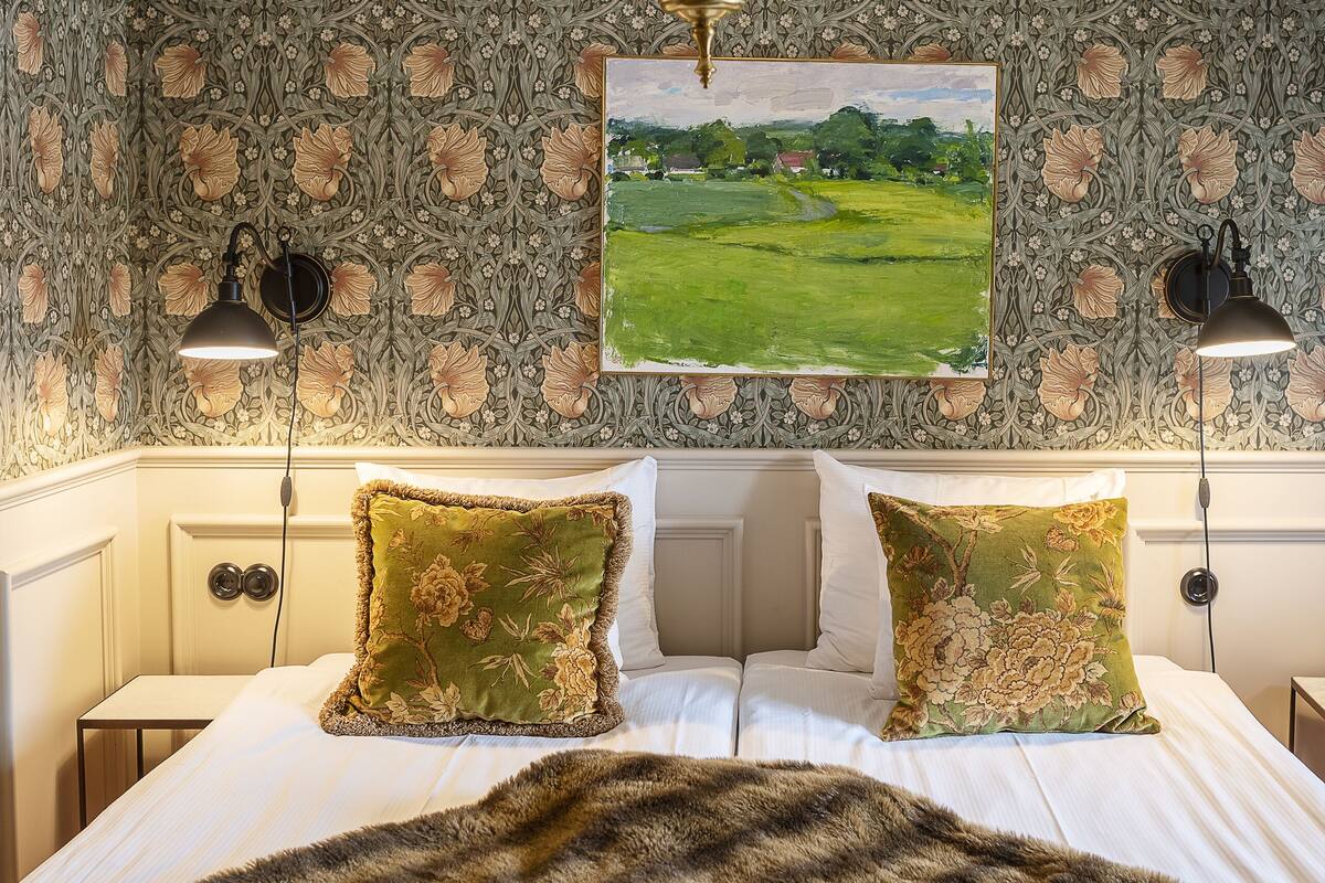 A bedroom with a large bed, floral patterned wallpaper, a painting of a landscape, and two bedside lamps.