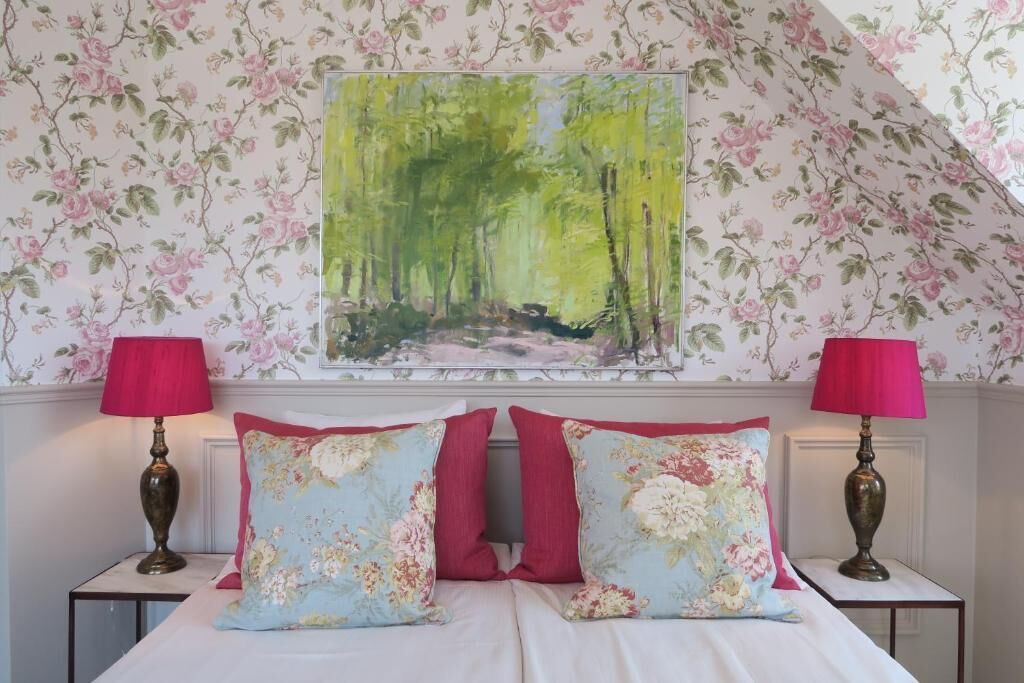 Un dormitorio con papel tapiz floral, un cuadro de un bosque y dos lámparas de noche.