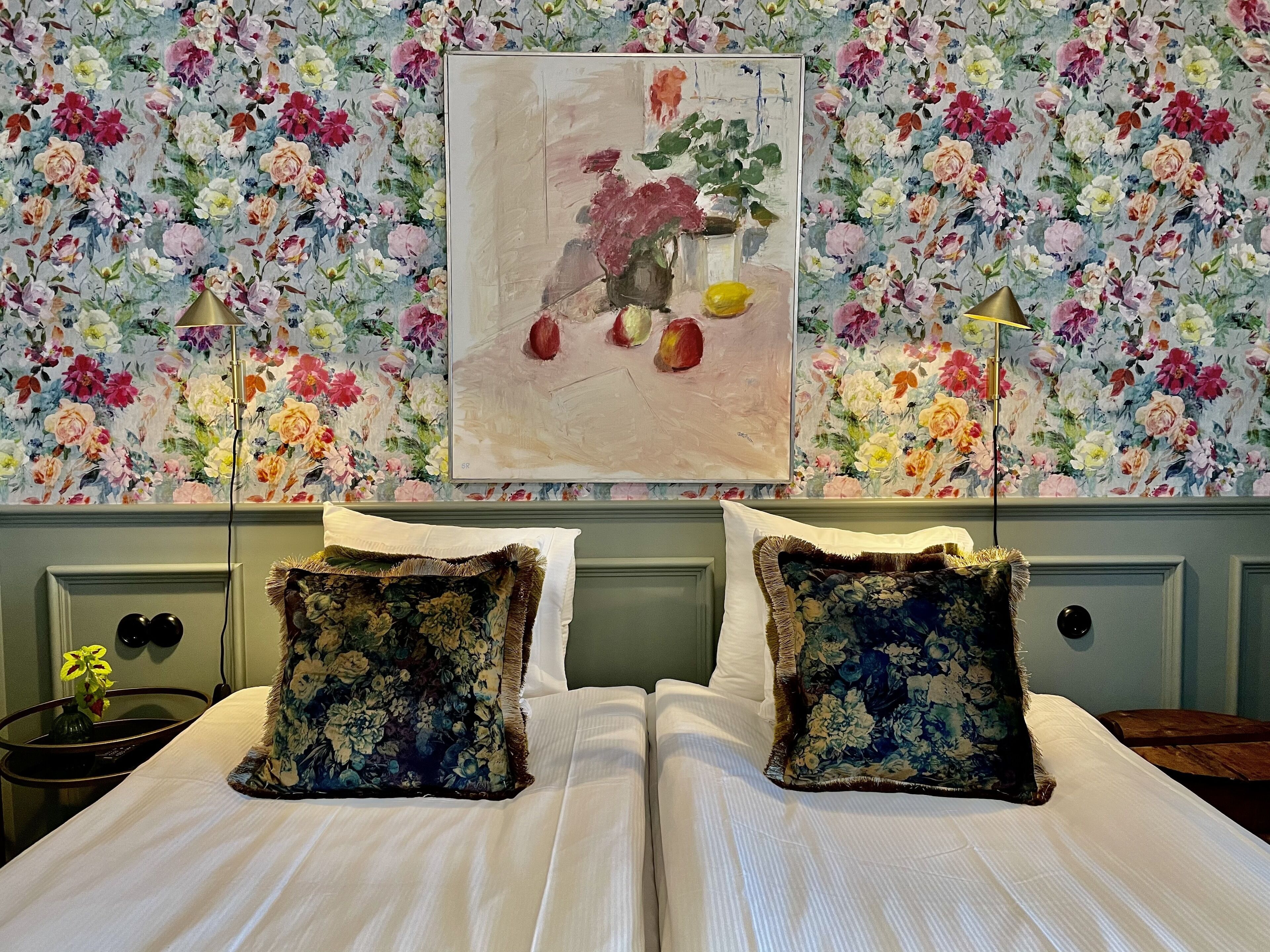 A hotel room with a floral wallpaper, a framed painting of flowers, two large floral pillows, and a bedside table with a lamp.