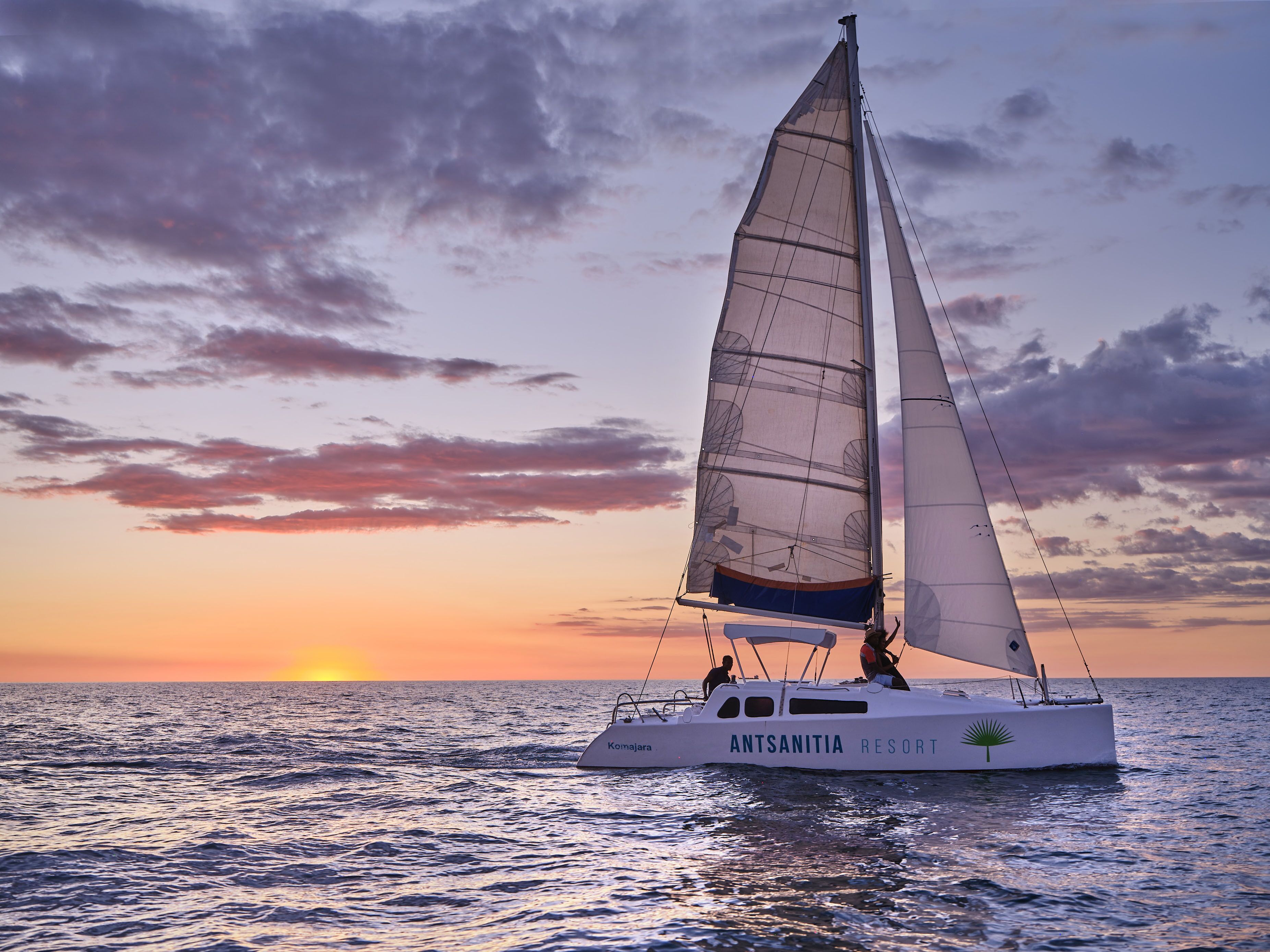 A sailboat named "Antsanitia Resort" with a person on deck and another person at the stern, sailing on calm waters during sunset.