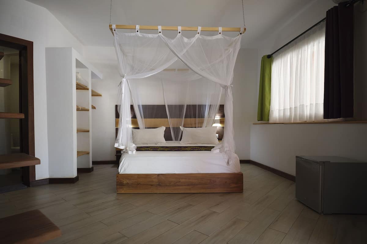 Un dormitorio con cama de madera, ropa de cama blanca, dosel, mesita de noche, armario y ventana con cortinas.