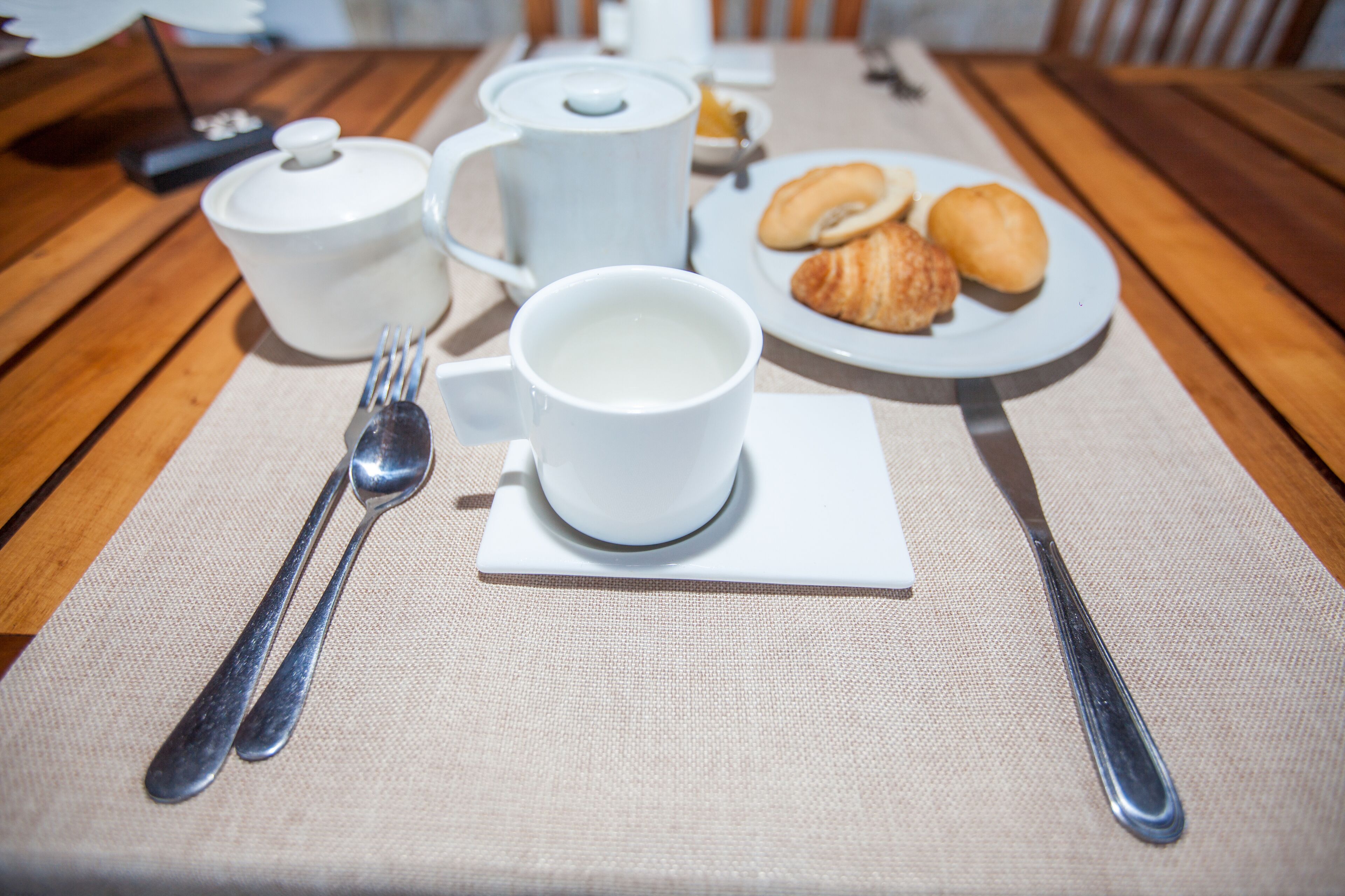 A breakfast setup with a teapot, cup, plate of croissants, and utensils on a wooden table.