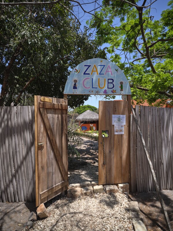 Una verja de madera con un letrero que dice "Zaza Club" y una estructura de techo de paja al fondo.