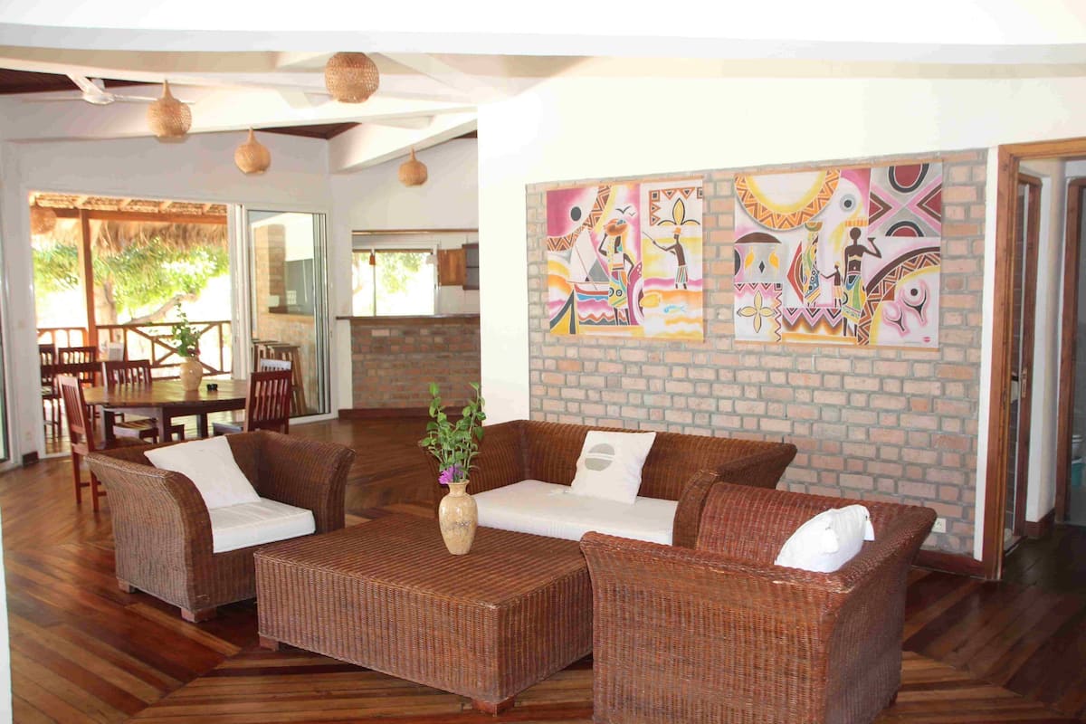 Amplia sala de estar con muebles de mimbre, un comedor y una pared de ladrillo con arte colorido.