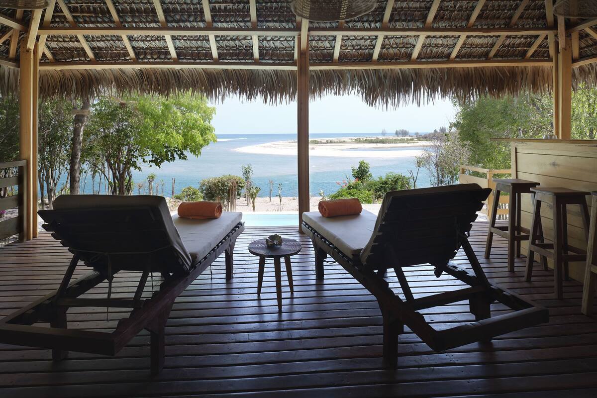 Una terraza de madera con dos sillas reclinatorias orientadas a vistas de la playa, una mesita y una estructura con techo de paja.